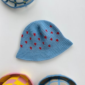 May include: A collection of crochet bucket hats in various colors. A light blue hat with red heart accents is the focal point. Other hats include a yellow and pink checkered hat, and hats with blue and white checkered patterns.