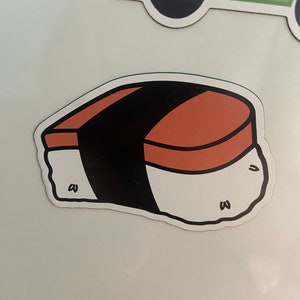 Puede incluir: Un imán con forma de sushi con una base blanca, una parte superior marrón y una franja negra en el medio. El imán tiene un borde blanco y pequeños detalles negros. El imán es probablemente para un refrigerador o una superficie similar.
