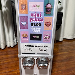 May include: A silver and white mini prints vending machine with colorful graphics. The machine features the text "mini Prints $1.00" and "2 quarters on each side". The graphics include hearts, food, and phrases.