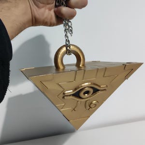 May include: A gold-colored, pyramid-shaped pendant with a chain. The pendant features an eye design and is held by a silver chain. The pendant is a unique accessory.