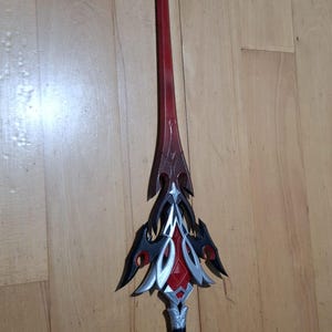 Może przedstawiać: Ozdobny miecz z czerwonym ostrzem i skomplikowanym srebrno-czarnym jelcem. Konstrukcja miecza charakteryzuje się spiczastą końcówką i ozdobnymi detalami, sugerując motyw fantasy lub cosplay. Miecz ma około 76 cm długości.