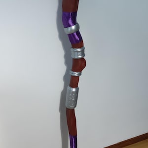 Pode incluir: Uma vara de madeira marrom com detalhes em roxo e prata. A vara tem uma ponta em forma de mão e é decorada com uma fita roxa e faixas prateadas.