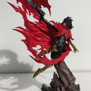 May include: A detailed figurine of a character in a dynamic pose, featuring a flowing red cape and black attire. The figure is holding a gun and is mounted on a brown, rocky base. The character has black hair and gold accents.
