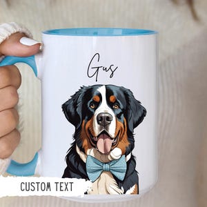May include: White ceramic mug with a blue rim and handle. The mug features a cartoon illustration of a Bernese Mountain Dog wearing a blue bow tie. The text "Gus" is printed on the mug in black cursive font. The text "CUSTOM TEXT" is printed on the bottom of the mug.