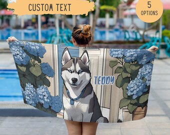Può includere: Un asciugamano da spiaggia con un'illustrazione di un cane husky di cartone animato. Il cane è seduto davanti a una porta con il nome "Teddy" scritto sopra. L'asciugamano ha un design floreale blu e verde.