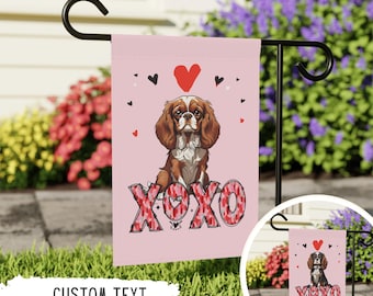May include: A pink garden flag with a cartoon Cavalier King Charles Spaniel dog wearing a pink and red heart pattern. The dog is sitting and the text "XOXO" is below the dog. The text is also in a pink and red heart pattern.