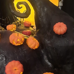 May include: A black and white figurine of Jack Skellington from The Nightmare Before Christmas, kissing a red-haired Sally figurine. The figurines are standing on a black, twisted, tree branch with a glowing orange moon in the background. There are several small orange pumpkins with glowing faces scattered around the base of the branch.