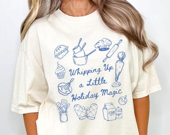 May include: Cream-colored t-shirt with a blue illustration of baking tools and the phrase "Whipping Up a Little Holiday Magic." The design includes a pie, cupcake, mixing bowls, chef's hat, rolling pin, and other baking-related icons.