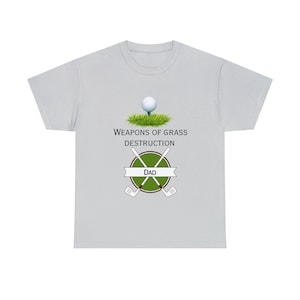 Puede incluir: Una camiseta gris claro con un diseño de temática de golf. El diseño presenta una bola de golf en un parche de hierba verde, palos de golf cruzados y el texto "Weapons of Grass Destruction" y "Dad".