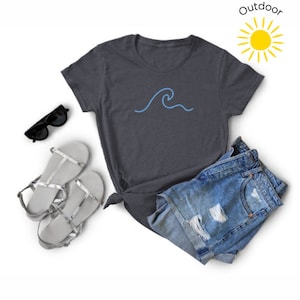 May include: A dark gray t-shirt with a blue wave graphic. The t-shirt is laid out on a white surface with a pair of silver sandals and denim shorts.