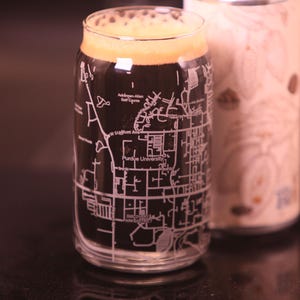 May include: A glass beer can-shaped glass with a map of Purdue University etched on the side, filled with dark beer and foam. A second, cylindrical container with a wood grain pattern is behind it.