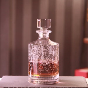 May include: A clear glass decanter with a stopper, filled with amber liquid. The decanter features a white etched map design. The decanter sits on a dark gray surface, with a blurred background.