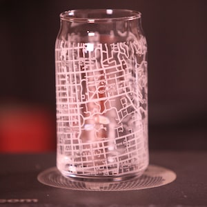 Puede incluir: Un vaso de beber de vidrio transparente con un diseño grabado en blanco de un mapa de la ciudad. El vaso tiene forma de lata y está sobre un posavasos.
