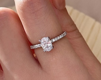 Peut inclure: Une bague en argent avec une pierre ovale transparente entourée de petites pierres transparentes. La bague est portée au doigt.