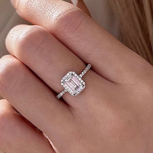 May include: A silver ring with a large, rectangular, clear gemstone surrounded by smaller clear gemstones. The ring is on a finger.