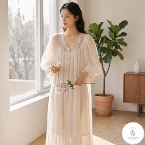 May include: A cream-coloured nightgown with lace detailing around the neckline and sleeves. The long-sleeved gown has a V-neck and a flowing, floor-length design. The model is holding flowers near a window with a potted plant in the background.