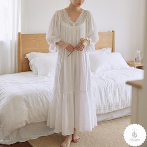 May include: A white nightdress with a V-neck and lace trim. The nightdress features long, puffed sleeves and a tiered skirt. A woman holds a dried yellow flower. The background shows a bed with white bedding and a wooden headboard.