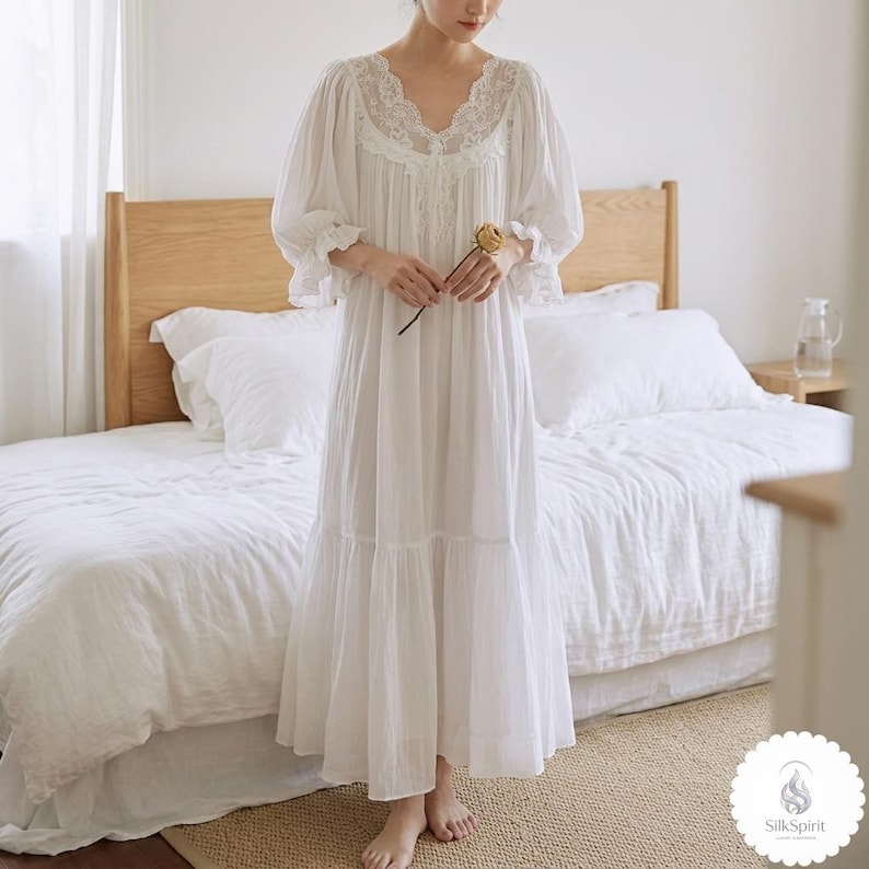 May include: A white nightdress with a V-neck and lace trim. The nightdress features long, puffed sleeves and a tiered skirt. A woman holds a dried yellow flower. The background shows a bed with white bedding and a wooden headboard.
