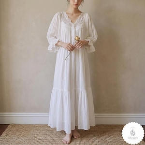 May include: A white nightdress with a V-neck and lace detailing. The nightdress features long sleeves with frilled cuffs and a tiered skirt. A person is holding a dried flower. The nightdress is made of a soft, flowing fabric.