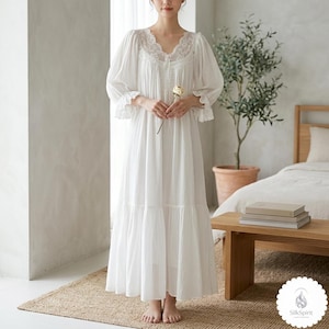 May include: A white nightdress with a lace-trimmed neckline and sleeves. The long gown has a tiered skirt and is made of a lightweight fabric. The model is holding a white rose. The image is set in a bedroom.