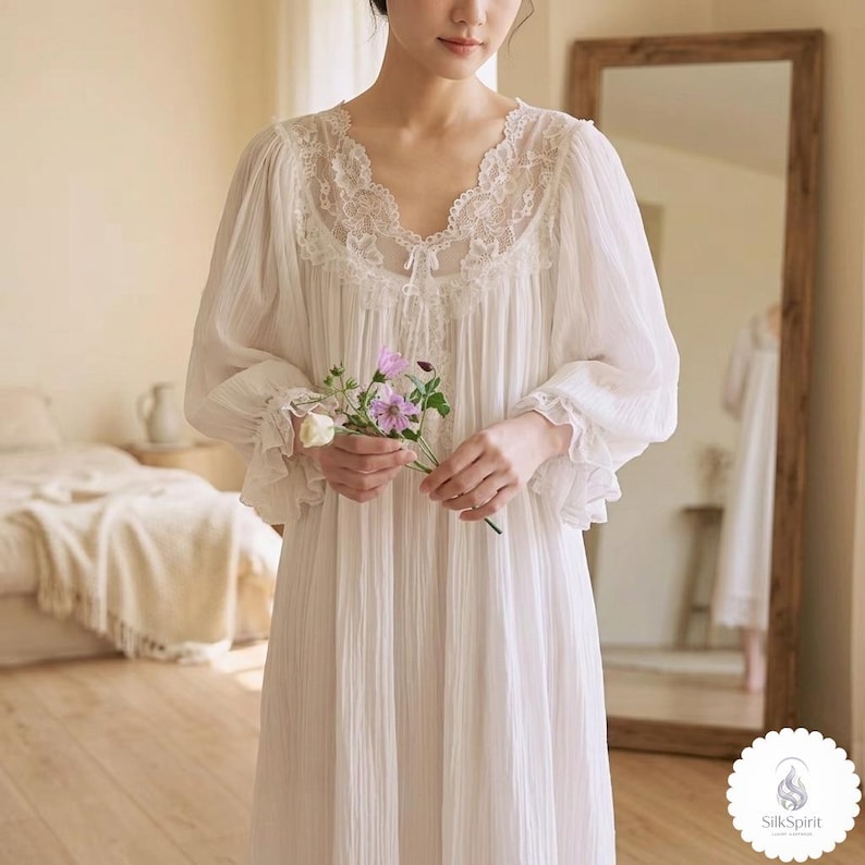 May include: A white nightdress with a V-neck and lace detailing. The long-sleeved gown has a pleated design and ruffled cuffs. The person is holding a small bouquet of purple and white flowers. The image is taken indoors.