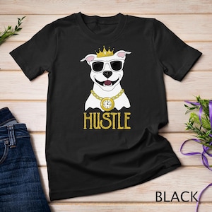 Puede incluir: Camiseta negra con un pitbull blanco que lleva una corona dorada, gafas de sol y una cadena de oro con un reloj de bolsillo. El texto "Hustle" está impreso en dorado debajo del perro.
