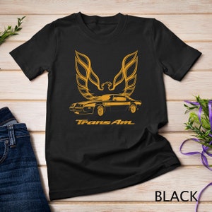 Puede incluir: Camiseta negra con un gráfico dorado de un coche clásico, el Pontiac Trans Am, con el icónico logotipo de pájaro encima. El texto "Trans Am" está impreso debajo del coche.