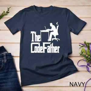 Könnte beinhalten: Ein marineblaues T-Shirt mit einer weißen Silhouette einer Person, die an einem Schreibtisch mit einem Laptop sitzt. Der Text "The CodeFather" ist in Weiß unter der Silhouette gedruckt.