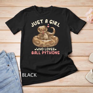 Puede incluir: Camiseta negra con el texto "Just a girl who loves ball pythons" en blanco. Un pitón bola de dibujos animados está enroscado en una cámara de aire rosa.