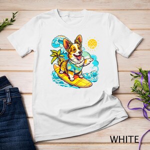 Puede incluir: Camiseta blanca con una imagen de dibujos animados de un perro corgi con una camisa floral y montando una tabla de surf en una ola. El perro sonríe y tiene una expresión feliz. El fondo es un océano azul con olas blancas y un sol amarillo.