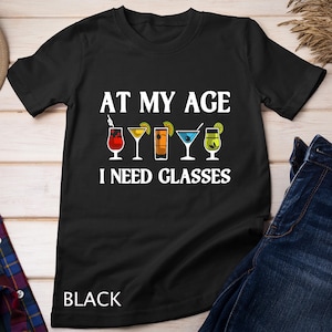 May include: Black t-shirt with the text "AT MY AGE I NEED GLASSES" above colorful cocktail illustrations. The word "BLACK" is printed at the bottom of the shirt. The shirt is made of a soft material.