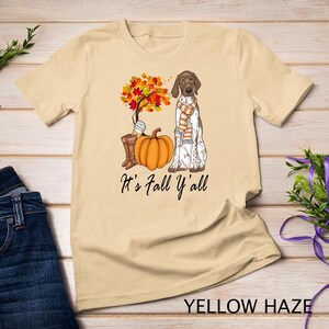 Könnte beinhalten: Ein beiges T-Shirt mit einem Aufdruck eines braunen und weißen Hundes, der mit einem Kürbis und Herbstblättern sitzt. Der Text "It's Fall Y'all" ist unter dem Aufdruck gedruckt.