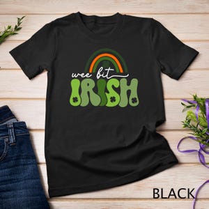 Puede incluir: Camiseta negra con un arcoíris verde y naranja y el texto "wee bit IRISH" en una fuente retro.