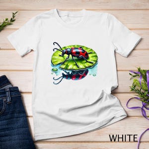 Könnte beinhalten: Weißes T-Shirt mit einem Aufdruck eines roten und schwarzen Käfers auf einem grünen Blatt. Der Käfer spiegelt sich im Wasser unter dem Blatt.