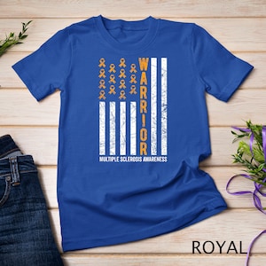 May include: A royal blue t-shirt with a distressed American flag design. The flag is white with orange ribbons in the top left corner. The word "WARRIOR" is written vertically in orange on the right side of the flag. The text "MULTIPLE SCLEROSIS AWARENESS" is written below the flag.