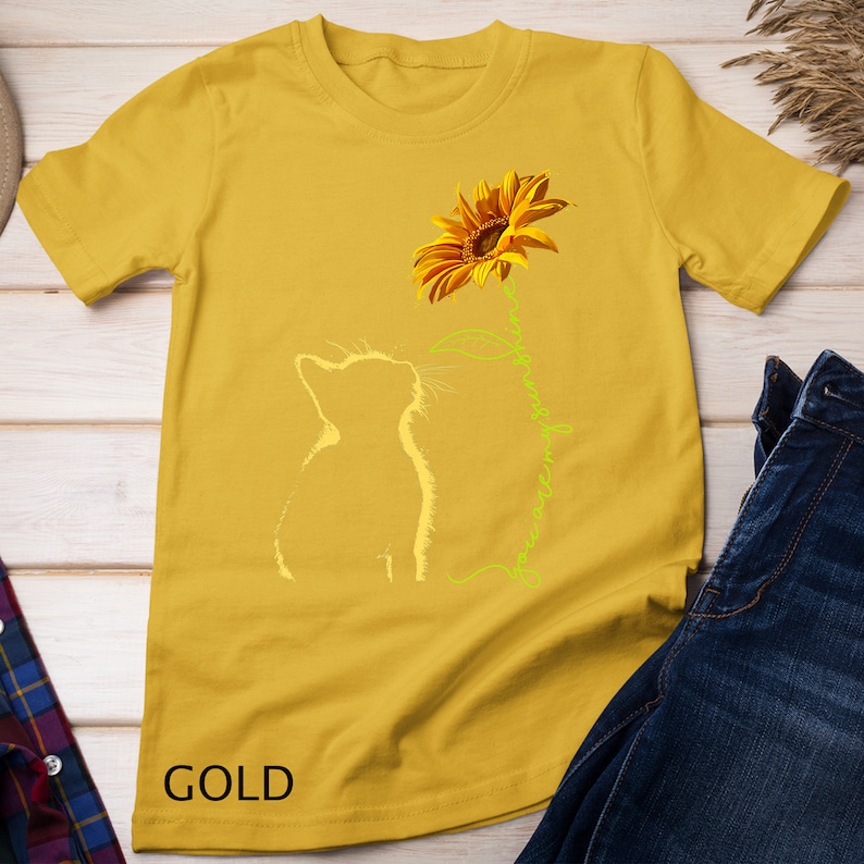 K&ouml;nnte beinhalten: Ein gelbes T-Shirt mit einer wei&szlig;en Silhouette einer Katze, die zu einer Sonnenblume aufblickt, mit dem Text "You are my sunshine" in Gr&uuml;n entlang des Stiels. Das T-Shirt liegt auf einer Holzoberfl&auml;che mit einer blauen Jeans und einem karierten Hemd.
