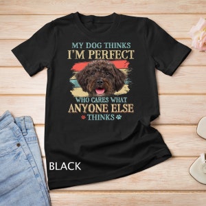 Może przedstawiać: Czarny T-shirt z grafiką w stylu vintage przedstawiającą brązowo-czarnego psa. Tekst na koszulce brzmi: "Mój pies myśli, że jestem idealny. Kogo obchodzi, co myślą inni."