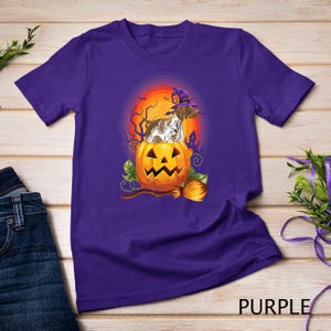 May include: Purple t-shirt featuring a graphic of a brown and white dog wearing a witch's hat sitting inside a jack-o'-lantern. The dog is surrounded by a broom and leaves.