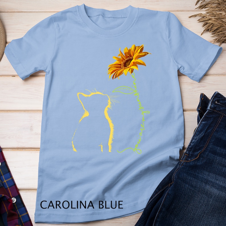 K&ouml;nnte beinhalten: Ein hellblaues T-Shirt mit einer gelben Sonnenblume und zwei gelben Katzen-Silhouetten. Der Text "You are my sunshine" ist in einer kursivschriftlichen Schriftart entlang des Stiels der Sonnenblume geschrieben.