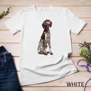 May include: White t-shirt featuring a brown and white German Shorthaired Pointer dog wearing red, white, and blue sunglasses. The dog is sitting and looking to the right.