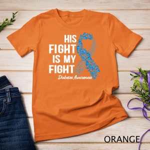 Puede incluir: Camiseta naranja con un gráfico blanco de una cinta azul hecha de mariposas. El texto en la camiseta dice "His Fight Is My Fight" y "Diabetes Awareness".