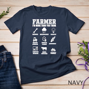 May include: A navy blue t-shirt with white text that reads "FARMER I'M MORE THAN YOU THINK". The shirt features a list of roles a farmer plays, including mechanic, meteorologist, scientist, machine operator, financial planner, agronomist, computer operator, animal caretaker, and family.
