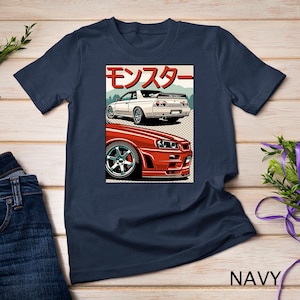 May include: A navy blue t-shirt with a graphic of two red and white Nissan Skyline GT-R cars. The graphic is in a retro style with the Japanese word "Monster" written in red on the top of the graphic.