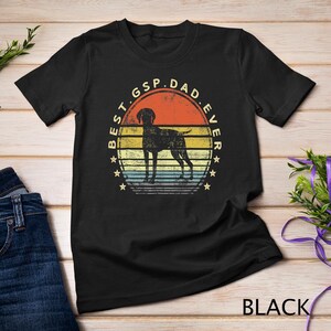 Pode incluir: Camiseta preta com um gráfico de estilo vintage de um cão Braco Alemão de Pelo Curto. O gráfico diz "Best GSP Dad Ever" com um fundo listrado vermelho, branco e azul.