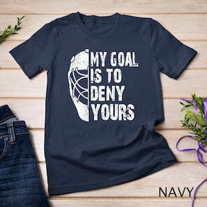 May include: Navy blue t-shirt featuring a white hockey goalie mask and the text "My goal is to deny yours".
