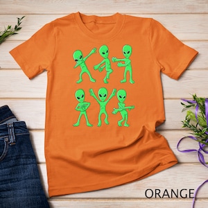 May include: Orange t-shirt featuring a graphic print of six green aliens dancing. The aliens are in different poses, some are doing the dab, others are waving their arms.