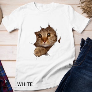 Op de afbeelding: Wit T-shirt met een afbeelding van een bruine tabbykat die door een gescheurd gat in de stof kijkt. De kat heeft grote groene ogen en een witte borst.