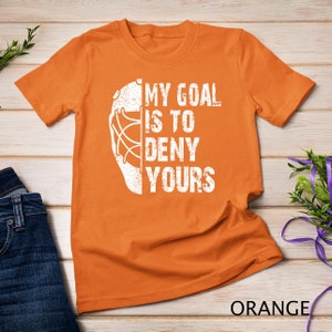 May include: Orange t-shirt with a white hockey goalie mask graphic and the text "My goal is to deny yours".