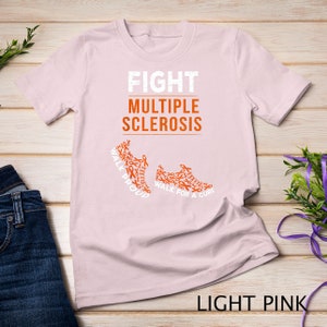 Op de afbeelding: Licht roze T-shirt met de tekst "FIGHT MULTIPLE SCLEROSIS" en twee hardloopschoenen gemaakt van oranje bewustzijn lintjes. De tekst op de schoenen luidt "WALK PROUD" en "WALK FOR A CURE".