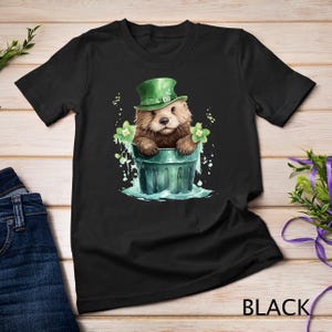 May include: A black t-shirt with a graphic of a cartoon otter wearing a green leprechaun hat and sitting in a green bucket. The otter is surrounded by green shamrocks.
